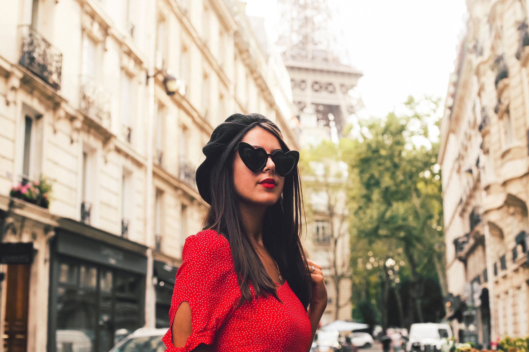 A woman in a beret, red dress and heart shaped sunglasses stands in a Paris street with the Eiffel Tower in the background. It's not Emily, but it has the same vibe.