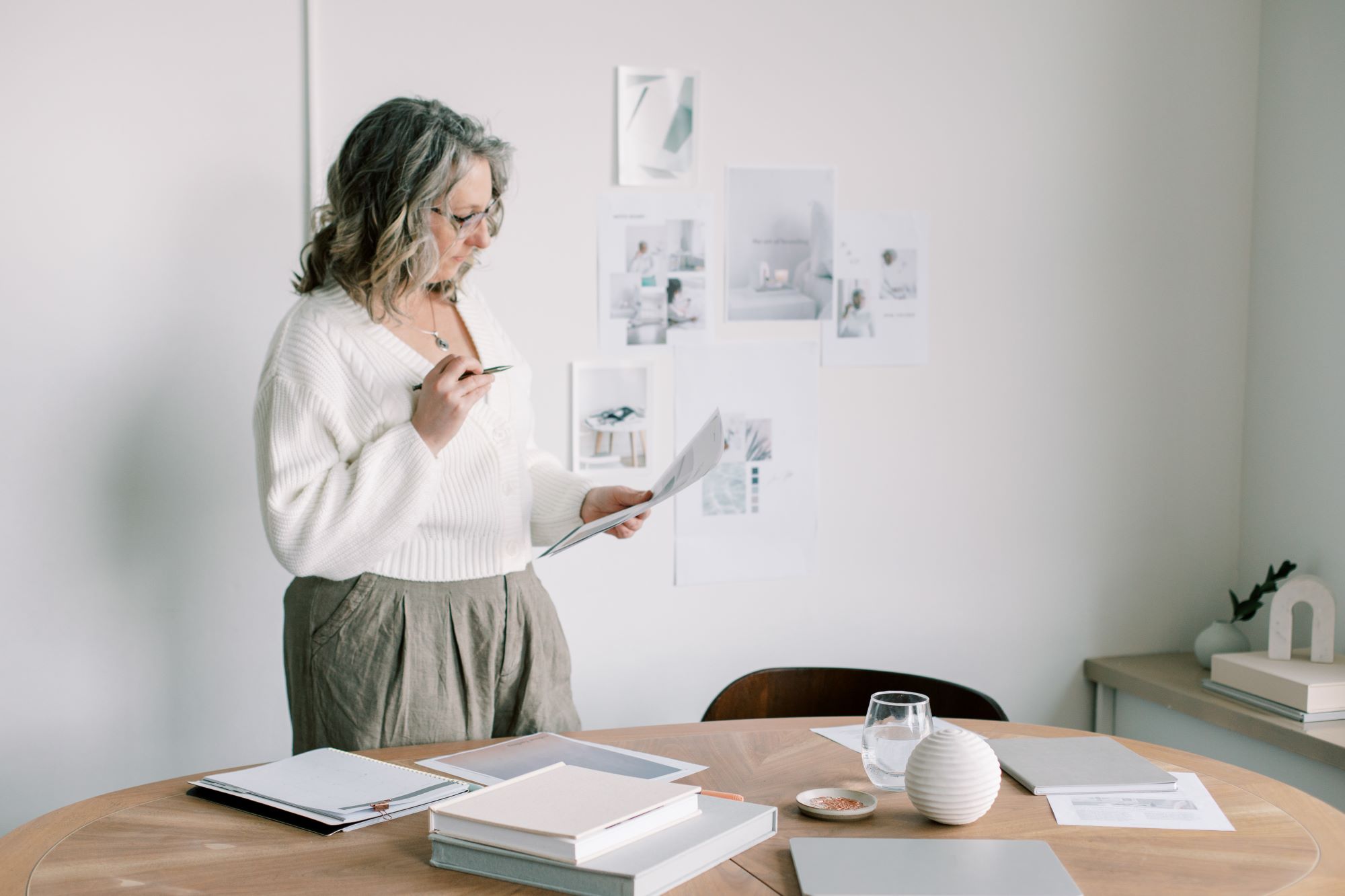 A woman stands behind a desk with a document in one hand and a pen in the other. The desk contains various reference materials and more are taped to the white wall behind her.